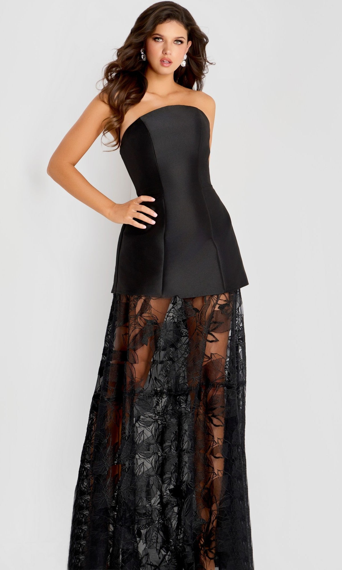 A woman poses with one hand on her hip in the Jovani 45467 long black prom dress, which features a strapless fitted bodice and a sheer lace skirt with floral patterns, set against a plain background.